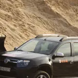 Czarna Dacia Duster 4x4 obok łyżki koparki na piaszczystym pustkowiu.