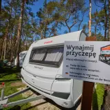 Biała przyczepa kempingowa nad morzem, otoczona sosnowym lasem. Tablica informuje o możliwości wynajmu.