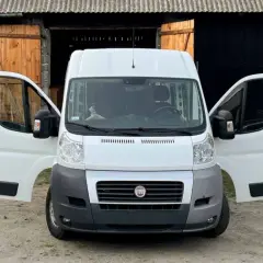 Biały bus z otwartymi drzwiami, stojący przed stodołą. Obok przyczepa i różne przedmioty, jakby ktoś zastanawiał się, jak nie budować kampera.