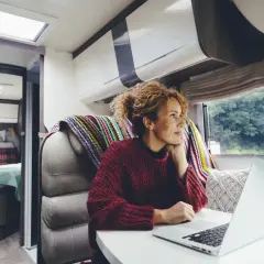 Kobieta z burzą loków, pracująca na laptopie w kamperze. Cieszy się wolnością, jaką daje życie cyfrowego nomady, z widokiem na zieleń za oknem.