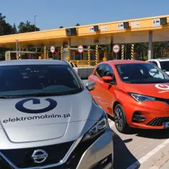 Srebrny Nissan i pomarańczowy Renault stoją przed bramkami poboru opłat. Na maskach aut widnieje logo "elektromobilni.pl". To idealne miejsce na podróż z przyczepą, nawet elektryczną!