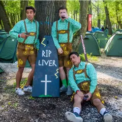 Trzech mężczyzn w strojach bawarskich przy nagrobku "RIP LIVER" na polu namiotowym. Prawdopodobnie uczestnicy imprezy w stylu Oktoberfest w Niemczech.