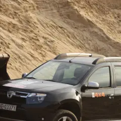 Czarna Dacia Duster 4x4 obok łyżki koparki na piaszczystym pustkowiu.