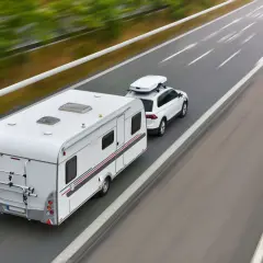Biały samochód z przyczepą kempingową i boxem dachowym jedzie autostradą. To podróż z wykorzystaniem viatoll, co ułatwia płatności za drogi.