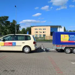 Samochód szkoleniowy z przyczepą, gotowy do nauki jazdy na przyczepę. Na drzwiach widnieje "L" i dane szkoły jazdy.