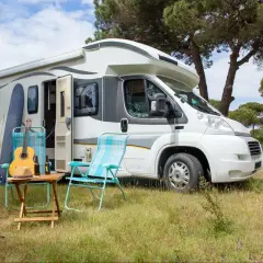 Idealny parking kamper: biały kamper pod drzewami, obok deska surfingowa, gitara, kapelusz i dwa krzesła.