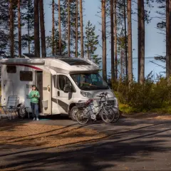 Kamper zaparkowany w lesie, obok jeziora. Na pierwszym planie dwa rowery, obok kampera siedzi osoba. Miejsce idealne na wypoczynek, pogoria 4 parking.