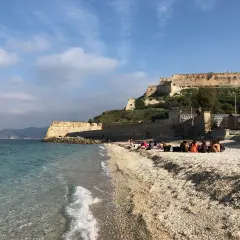 Plaża na wyspie Elba we Włoszech, z turkusową wodą i kamienistym brzegiem. W tle widać zabytkową fortecę na wzgórzu.