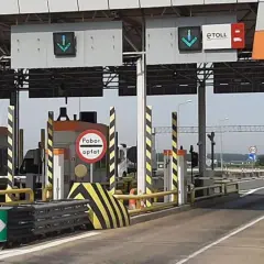 Biały samochód z przyczepą zbliża się do punktu poboru opłat za przejazd autostradą. Widać bramki i znaki informujące o opłatach.