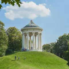 Monachijski ogród angielski z Monopterosem na wzgórzu. Ludzie spacerują i jeżdżą na rowerach po zielonych trawnikach.