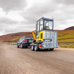 Czarny SUV ciągnie na przyczepie lekkiej żółtą koparkę JCB. Pojazd z przyczepą jedzie po pustej drodze wśród pagórków.