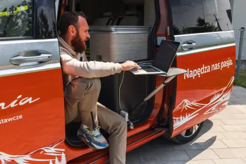 Mężczyzna pracuje na laptopie przy otwartych drzwiach kampera. Idealne miejsce na podróż kamperem, by połączyć pracę z przygodą.