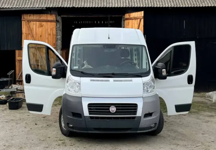 Biały bus z otwartymi drzwiami, stojący przed stodołą. Obok przyczepa i różne przedmioty, jakby ktoś zastanawiał się, jak nie budować kampera.