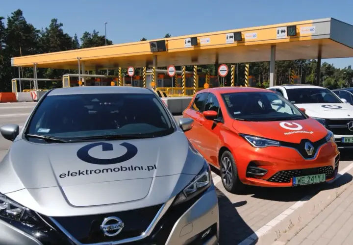 Srebrny Nissan i pomarańczowy Renault stoją przed bramkami poboru opłat. Na maskach aut widnieje logo "elektromobilni.pl". To idealne miejsce na podróż z przyczepą, nawet elektryczną!