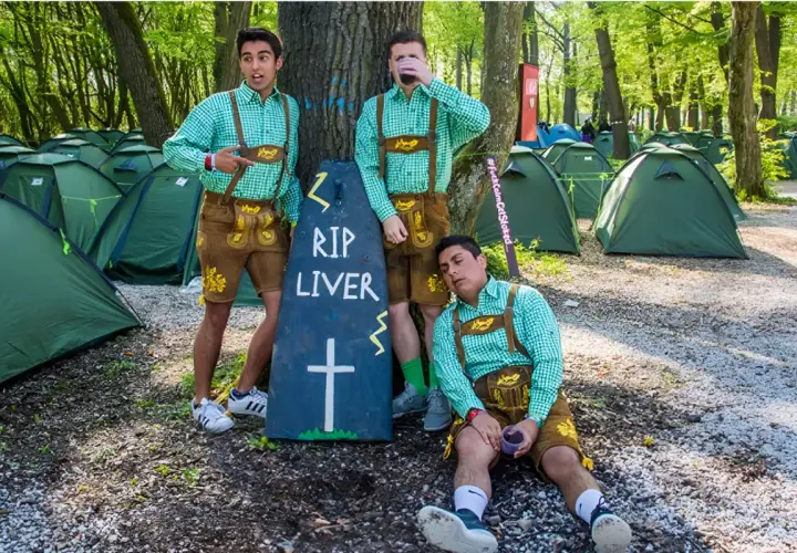 Trzech mężczyzn w strojach bawarskich przy nagrobku "RIP LIVER" na polu namiotowym. Prawdopodobnie uczestnicy imprezy w stylu Oktoberfest w Niemczech.