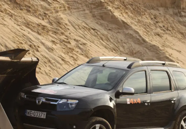 Czarna Dacia Duster 4x4 obok łyżki koparki na piaszczystym pustkowiu.
