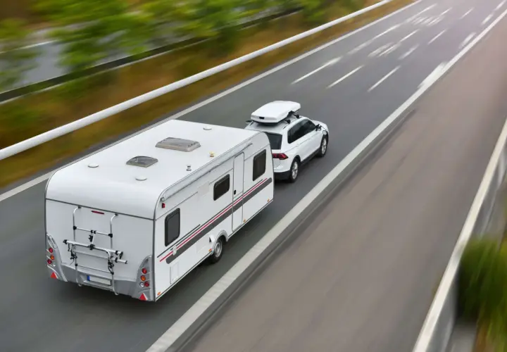 Biały samochód z przyczepą kempingową i boxem dachowym jedzie autostradą. To podróż z wykorzystaniem viatoll, co ułatwia płatności za drogi.