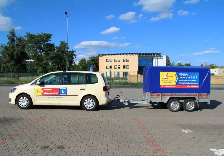 Samochód szkoleniowy z przyczepą, gotowy do nauki jazdy na przyczepę. Na drzwiach widnieje "L" i dane szkoły jazdy.