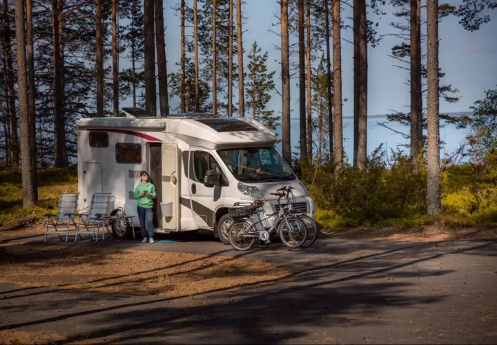 Kamper zaparkowany w lesie, obok jeziora. Na pierwszym planie dwa rowery, obok kampera siedzi osoba. Miejsce idealne na wypoczynek, pogoria 4 parking.