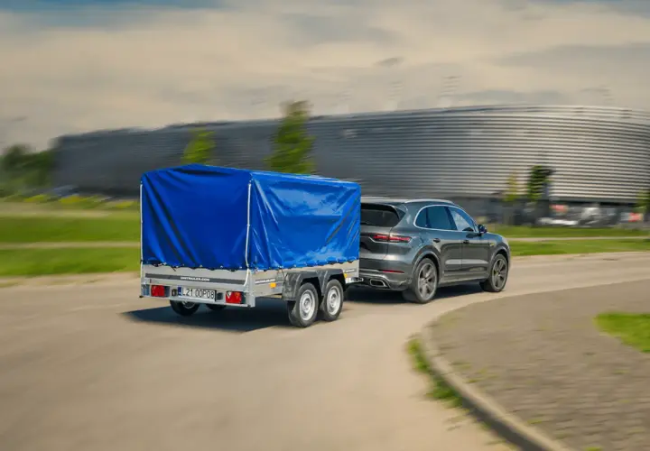 Szare Porsche Cayenne z niebieską plandeką na przyczepie dwuosiowej. Czy można ciągnąć przyczepę dwuosiową na kat. B?