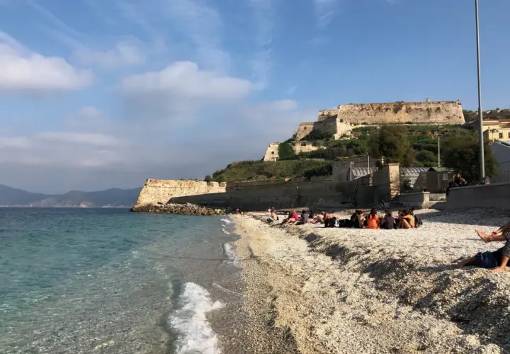 Plaża na wyspie Elba we Włoszech, z turkusową wodą i kamienistym brzegiem. W tle widać zabytkową fortecę na wzgórzu.