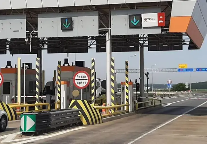 Biały samochód z przyczepą zbliża się do punktu poboru opłat za przejazd autostradą. Widać bramki i znaki informujące o opłatach.