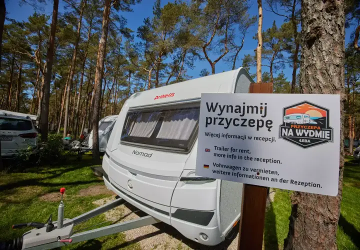 Biała przyczepa kempingowa nad morzem, otoczona sosnowym lasem. Tablica informuje o możliwości wynajmu.