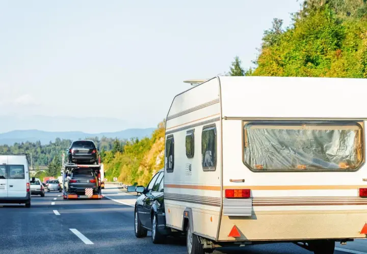 Samochód ciągnie przyczepę kempingową. Na autostradzie widać też inne pojazdy, w tym lawetę z samochodami. Winieta co to? To opłata za przejazd.