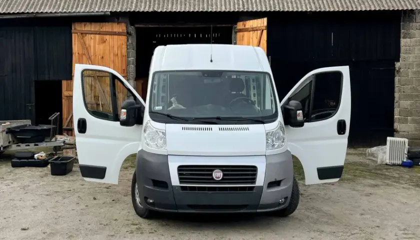 Biały bus z otwartymi drzwiami, stojący przed stodołą. Obok przyczepa i różne przedmioty, jakby ktoś zastanawiał się, jak nie budować kampera.