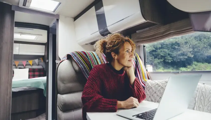 Kobieta z burzą loków, pracująca na laptopie w kamperze. Cieszy się wolnością, jaką daje życie cyfrowego nomady, z widokiem na zieleń za oknem.