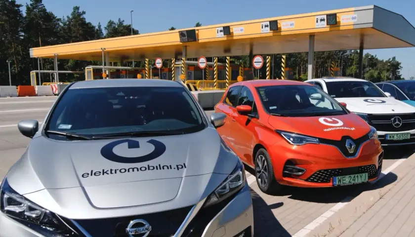 Srebrny Nissan i pomarańczowy Renault stoją przed bramkami poboru opłat. Na maskach aut widnieje logo "elektromobilni.pl". To idealne miejsce na podróż z przyczepą, nawet elektryczną!