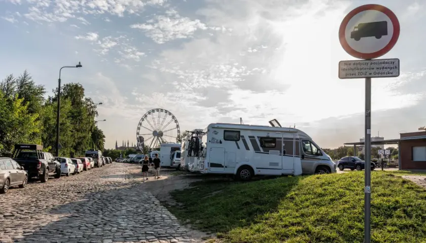 Darmowe parkingi Łeba: kamienista droga, kampery, samochody i diabelski młyn w tle.
