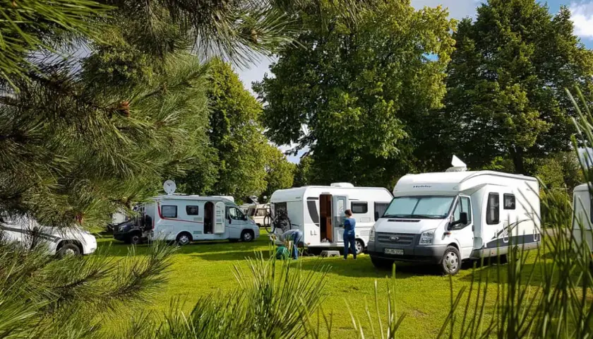 Kampery i przyczepy kempingowe na zielonej trawie, wśród drzew. Słoneczny dzień w **nude camp**.