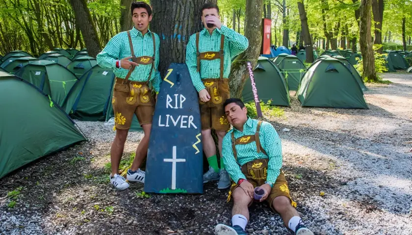 Trzech mężczyzn w strojach bawarskich przy nagrobku "RIP LIVER" na polu namiotowym. Prawdopodobnie uczestnicy imprezy w stylu Oktoberfest w Niemczech.