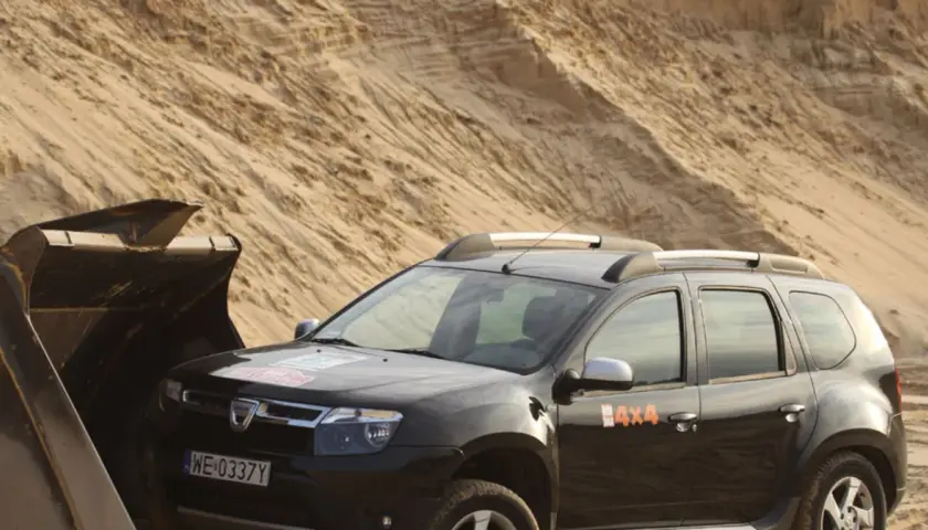 Czarna Dacia Duster 4x4 obok łyżki koparki na piaszczystym pustkowiu.