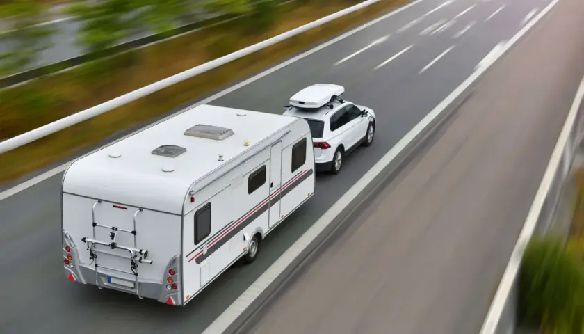 Biały samochód z przyczepą kempingową i boxem dachowym jedzie autostradą. To podróż z wykorzystaniem viatoll, co ułatwia płatności za drogi.