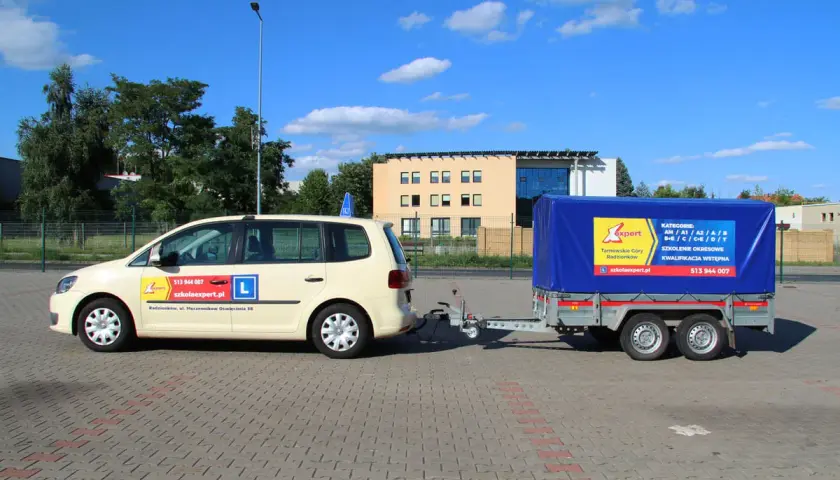 Samochód szkoleniowy z przyczepą, gotowy do nauki jazdy na przyczepę. Na drzwiach widnieje "L" i dane szkoły jazdy.