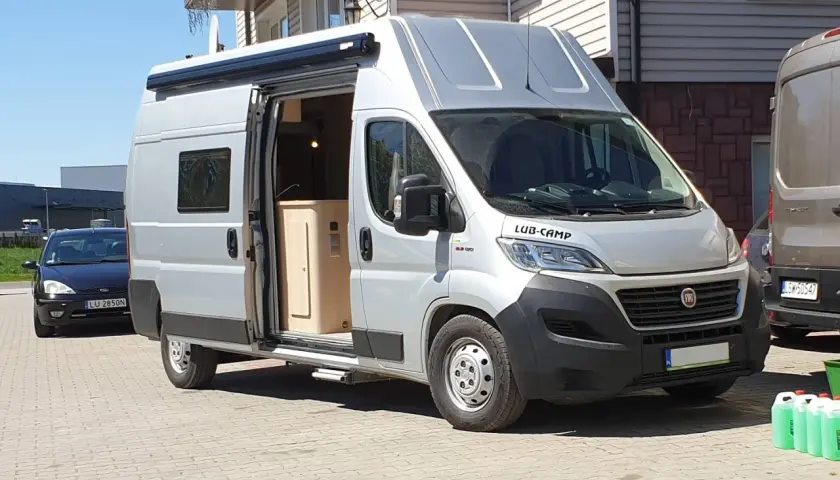 Srebrny kamper Ducato L3H3, z otwartymi drzwiami, ukazuje wnętrze. Idealny do podróży, jego wymiary pozwalają na komfort.
