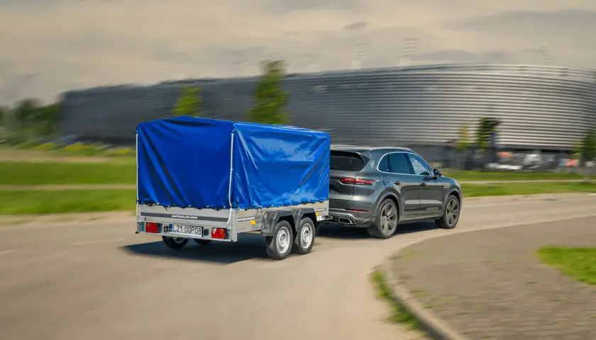 Szare Porsche Cayenne z niebieską plandeką na przyczepie dwuosiowej. Czy można ciągnąć przyczepę dwuosiową na kat. B?