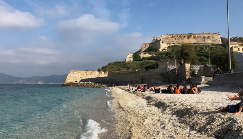Plaża na wyspie Elba we Włoszech, z turkusową wodą i kamienistym brzegiem. W tle widać zabytkową fortecę na wzgórzu.