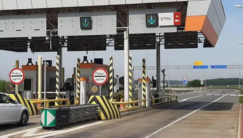 Biały samochód z przyczepą zbliża się do punktu poboru opłat za przejazd autostradą. Widać bramki i znaki informujące o opłatach.