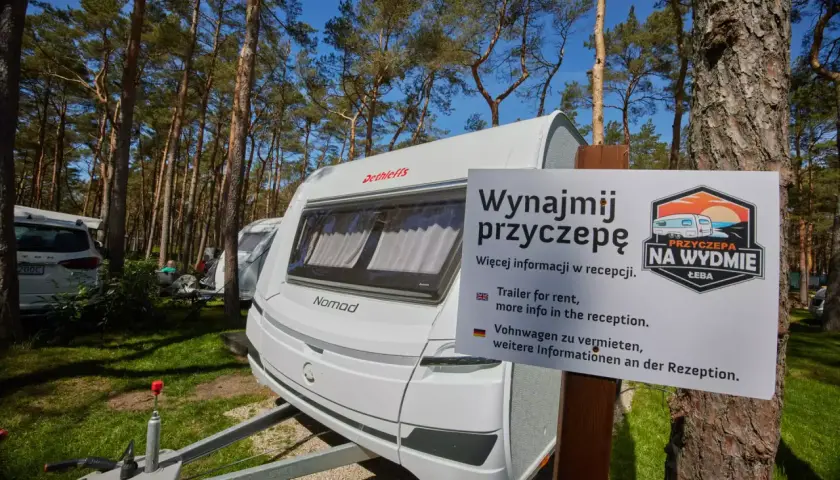 Biała przyczepa kempingowa nad morzem, otoczona sosnowym lasem. Tablica informuje o możliwości wynajmu.