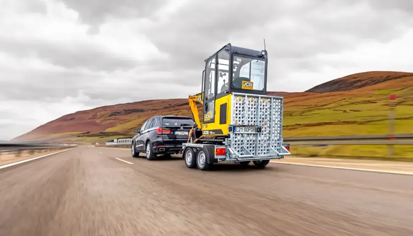 Czarny SUV ciągnie na przyczepie lekkiej żółtą koparkę JCB. Pojazd z przyczepą jedzie po pustej drodze wśród pagórków.