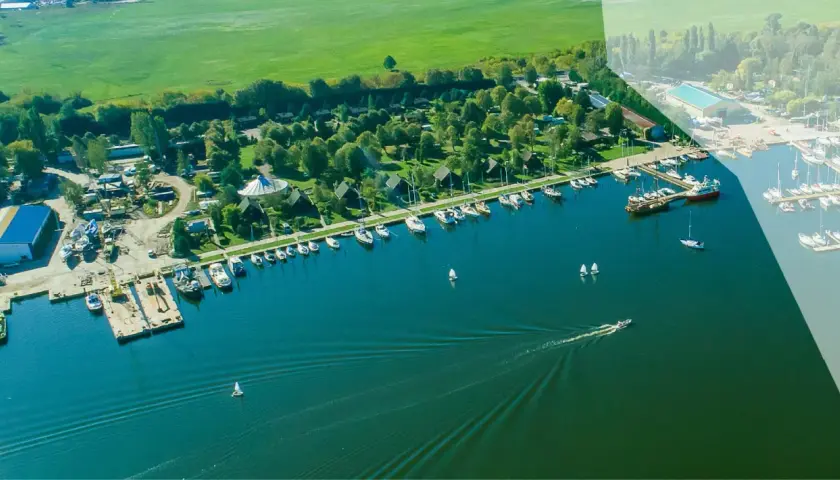 Widok z lotu ptaka na camping marina PTTK z licznymi łodziami i żaglówkami zacumowanymi przy brzegu, otoczony zielenią i polami.
