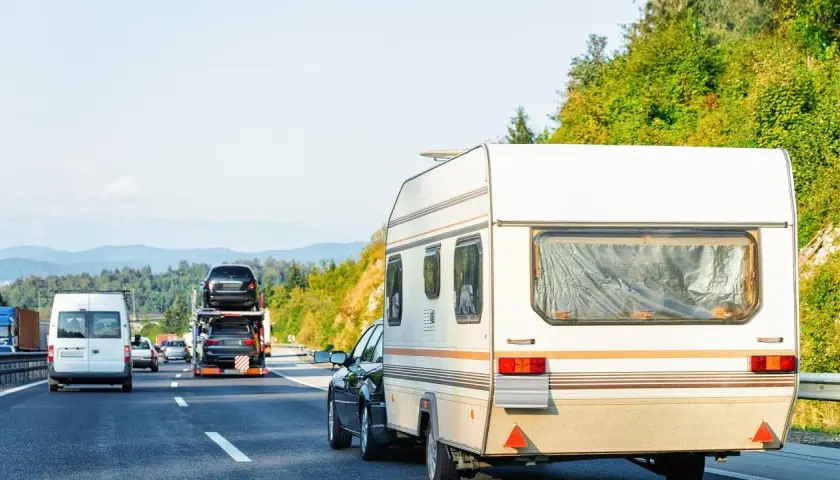 Samochód ciągnie przyczepę kempingową. Na autostradzie widać też inne pojazdy, w tym lawetę z samochodami. Winieta co to? To opłata za przejazd.