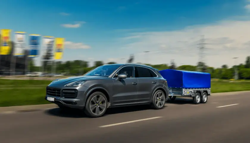 Szary SUV z przyczepą pod niebieską plandeką toczy się po autostradzie.