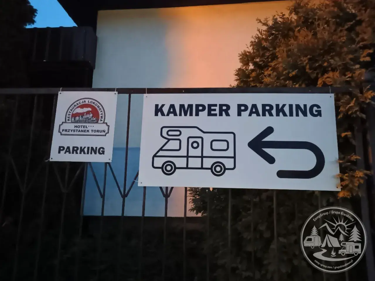 Znak "KAMPER PARKING" z ikoną kampera i strzałką w lewo. Obok znak "PARKING" z logo restauracji i hotelu.