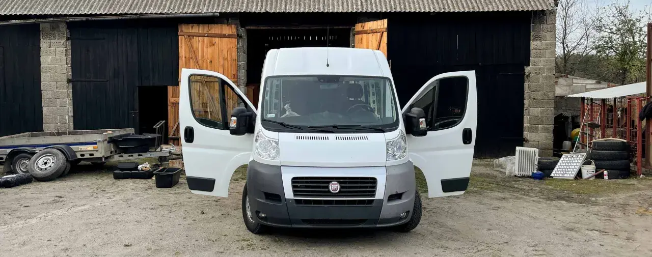Biały bus z otwartymi drzwiami, stojący przed stodołą. Obok przyczepa i różne przedmioty, jakby ktoś zastanawiał się, jak nie budować kampera.