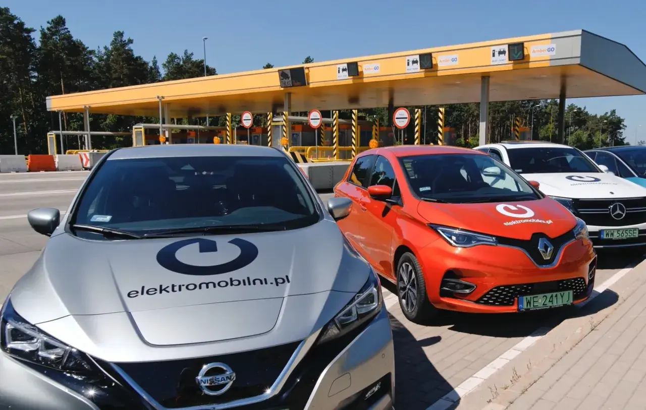 Srebrny Nissan i pomarańczowy Renault stoją przed bramkami poboru opłat. Na maskach aut widnieje logo "elektromobilni.pl". To idealne miejsce na podróż z przyczepą, nawet elektryczną!