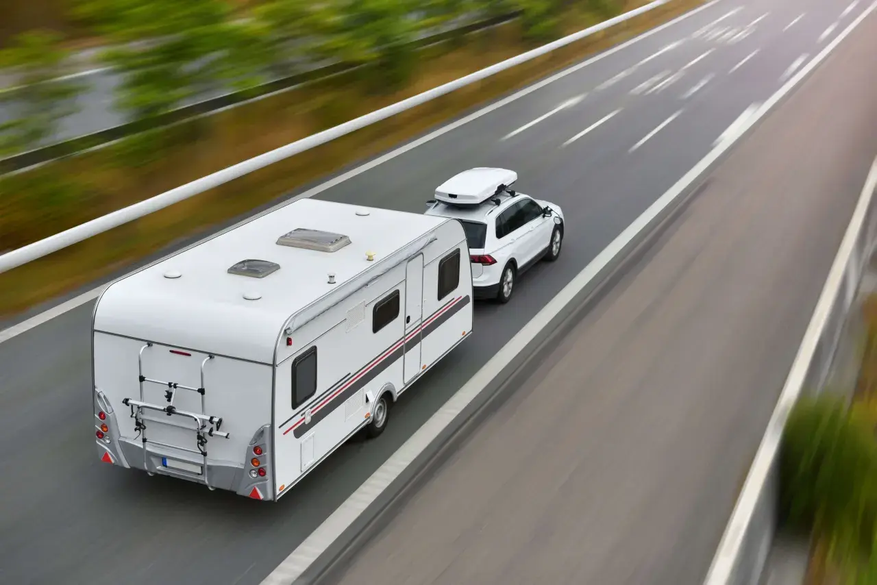 Biały samochód z przyczepą kempingową i boxem dachowym jedzie autostradą. To podróż z wykorzystaniem viatoll, co ułatwia płatności za drogi.